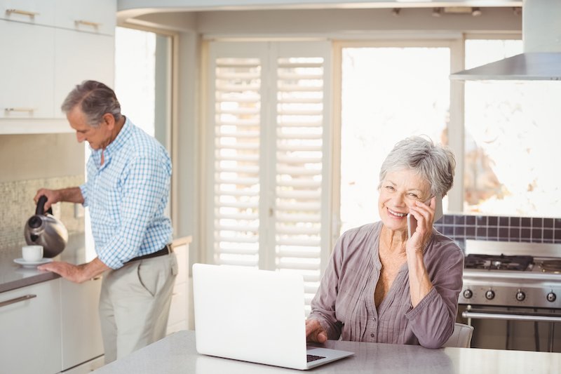 Senior woman talking on mobile phone with husband making tea in background at home